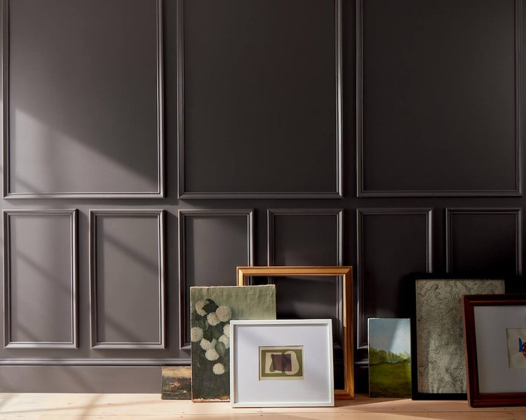 Artwork and frames arranged on a floor against a deep chocolate brown-painted wall with decorative molding.